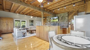 A spacious living area with a wooden ceiling, a stone fireplace, a kitchen with a stone wall, and a dining area with a marble table. - Mountain View Cabin in VA Wine Country (Scottsville)