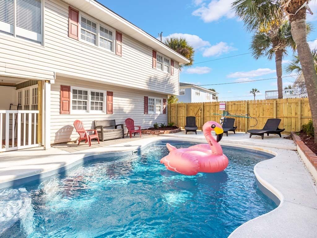 Uma piscina com um flutuador em formato de flamingo rosa, cercada por uma cerca de madeira, e uma casa com revestimento branco e persianas vermelhas ao fundo.