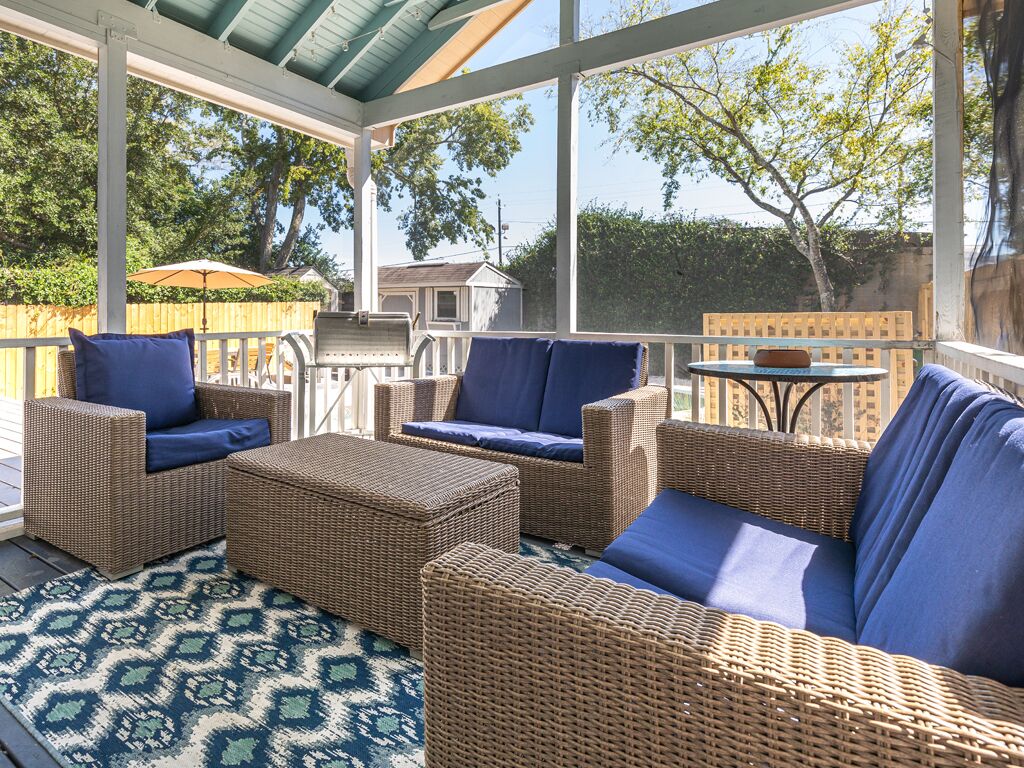 A covered outdoor seating area with wicker furniture and blue cushions, a patterned rug, and a view of the backyard.