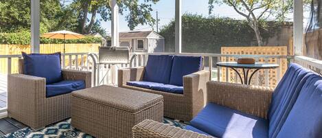 A covered outdoor seating area with wicker furniture and blue cushions, a patterned rug, and a view of the backyard.