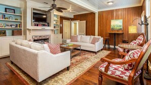 A living room with a fireplace, sofa, and armchairs. There is a bookshelf and a painting on the wall. - Frogtown Cottage by Tybee Vacation Rentals (Savannah)