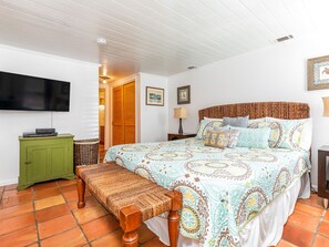 A bedroom with a large bed, a green nightstand, a television mounted on the wall, and a wooden bench. - Guiding Light Cottage by Tybee Vacation Rentals (Tybee Island)