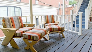 A covered outdoor deck with two striped armchairs and a small table, a staircase, and a brick building in the background.