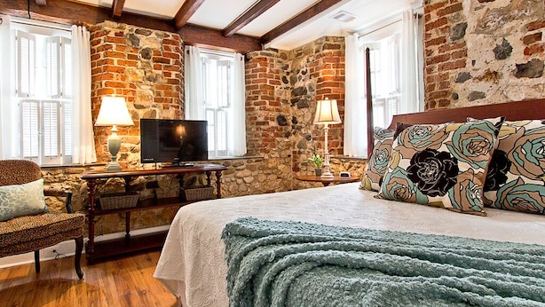 A room with exposed brick walls, wooden beams, a bed with floral pillows, a television, and a lamp.