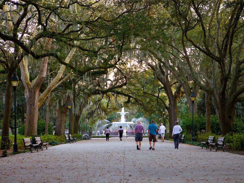 A park with a central fountain, benches, and walking paths lined with trees.