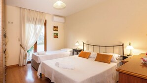 A hotel room with two beds, a wooden floor, a window with curtains, a bedside table with a lamp, and a wall-mounted air conditioner.