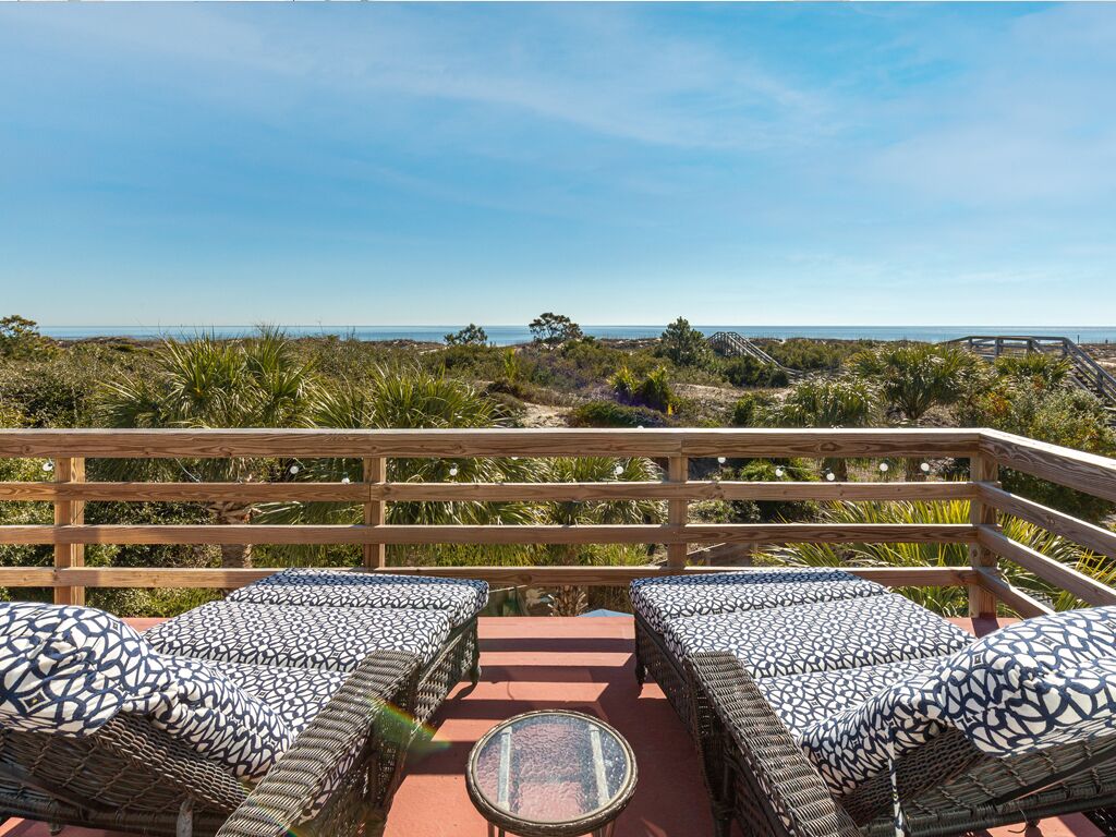 A balcony with wicker furniture and a glass table, overlooking a beach and ocean.
