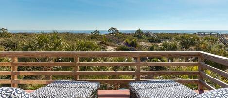 A balcony with wicker furniture and a glass table, overlooking a beach and ocean.