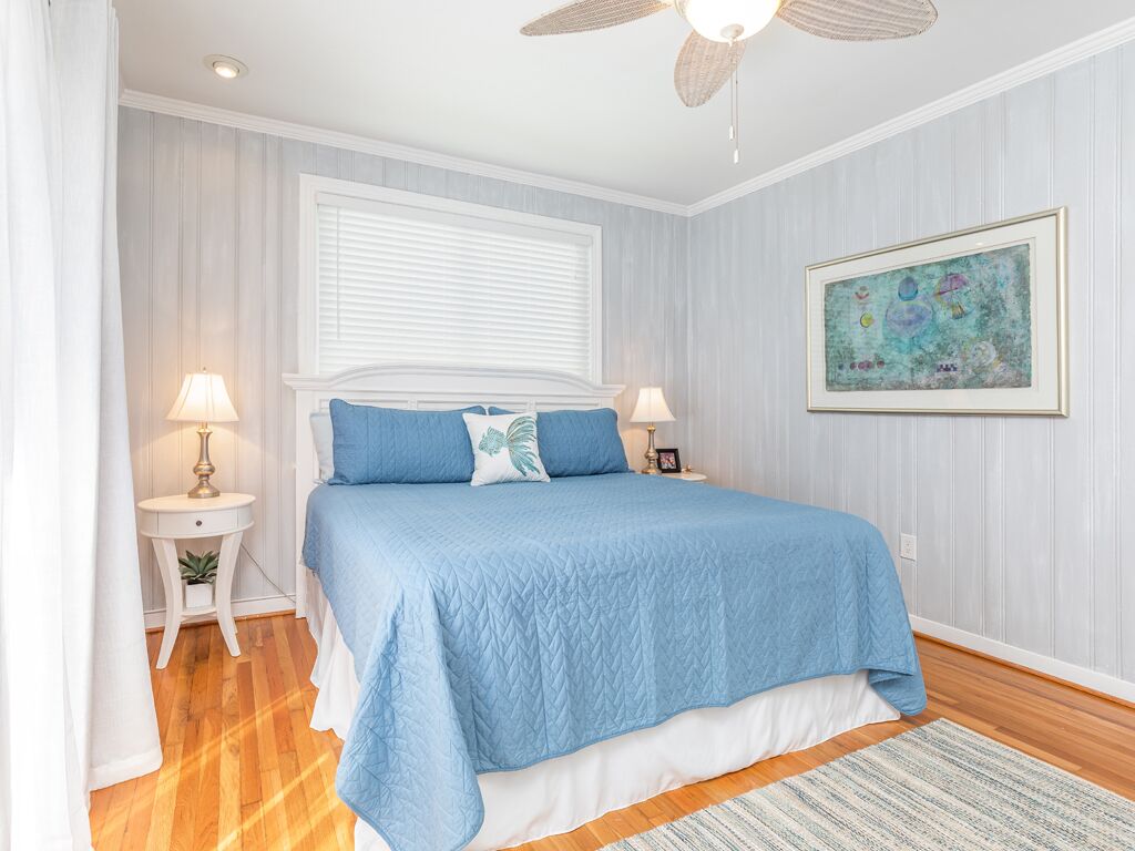 Une chambre avec un grand lit, deux tables de chevet, un ventilateur de plafond, une fenêtre avec des stores et une œuvre d’art encadrée accrochée au mur.