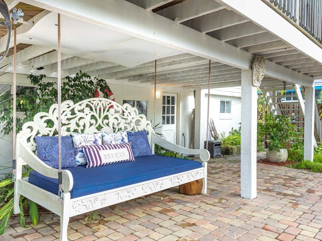 A white swing bed with blue cushions and a decorative headboard, set on a brick patio with plants and a white railing in the background.