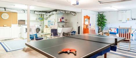 A ping pong table in a room with a washing machine, a fan, and a plant.