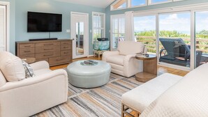 A spacious room with a flat-screen TV, a ceiling fan, a large window with a view, a sofa, a chair, and a bed. - Hang Ten by Tybee Vacation Rentals (Tybee Island)