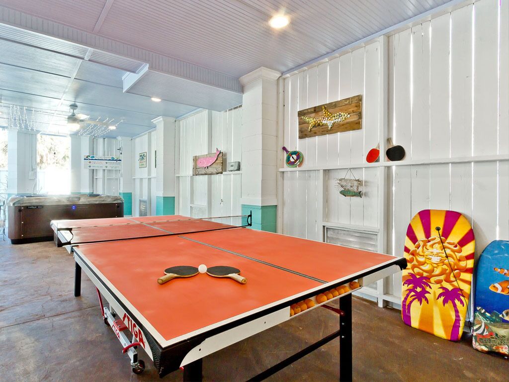 A ping pong table in a room with white paneled walls, a wooden ceiling, and various wall decorations including a shark, a fish, and a colorful surfboard.