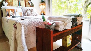 A neatly made bed with a floral-patterned blanket, a bedside table with a lamp, and a window with a view of trees. - Mountain house with panoramic viewpoint.  (Florianópolis)