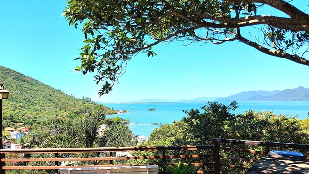 A scenic view of a coastal area with a wooden deck, potted plants, and a clear blue sky. - Mountain house with panoramic viewpoint.  (Florianópolis)