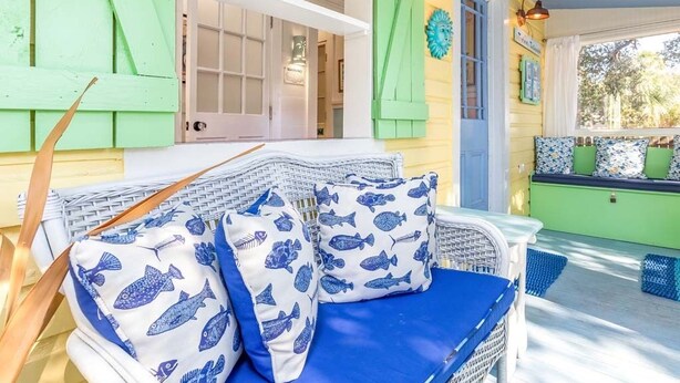 A bright, colorful porch with a wicker sofa and cushions, a green door, and a window with a view of the outdoors. - Island Bliss Tybee Vacation Rentals (Tybee Island)