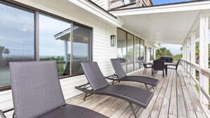 A covered outdoor deck with wicker lounge chairs, a grill, and large windows offering a view of the ocean.