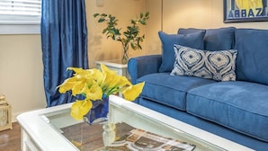 Un salon avec un canapé bleu, une table basse en verre sur laquelle repose un magazine, des fleurs jaunes et une lampe.