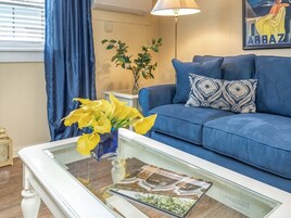 Un salón con un sofá azul, una mesa de centro de cristal con una revista, flores amarillas y una lámpara.