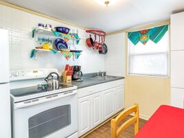 Una cocina compacta con armarios blancos, estufa y pileta. Se observan vajilla y utensilios de colores en las repisas. Se ve una ventana con cortina.