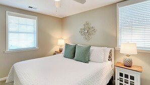 A bedroom with a large bed, two bedside tables with lamps, a ceiling fan, and a decorative wall piece. - Latitude Adjustment A Tybee Vacation Rentals (Tybee Island)