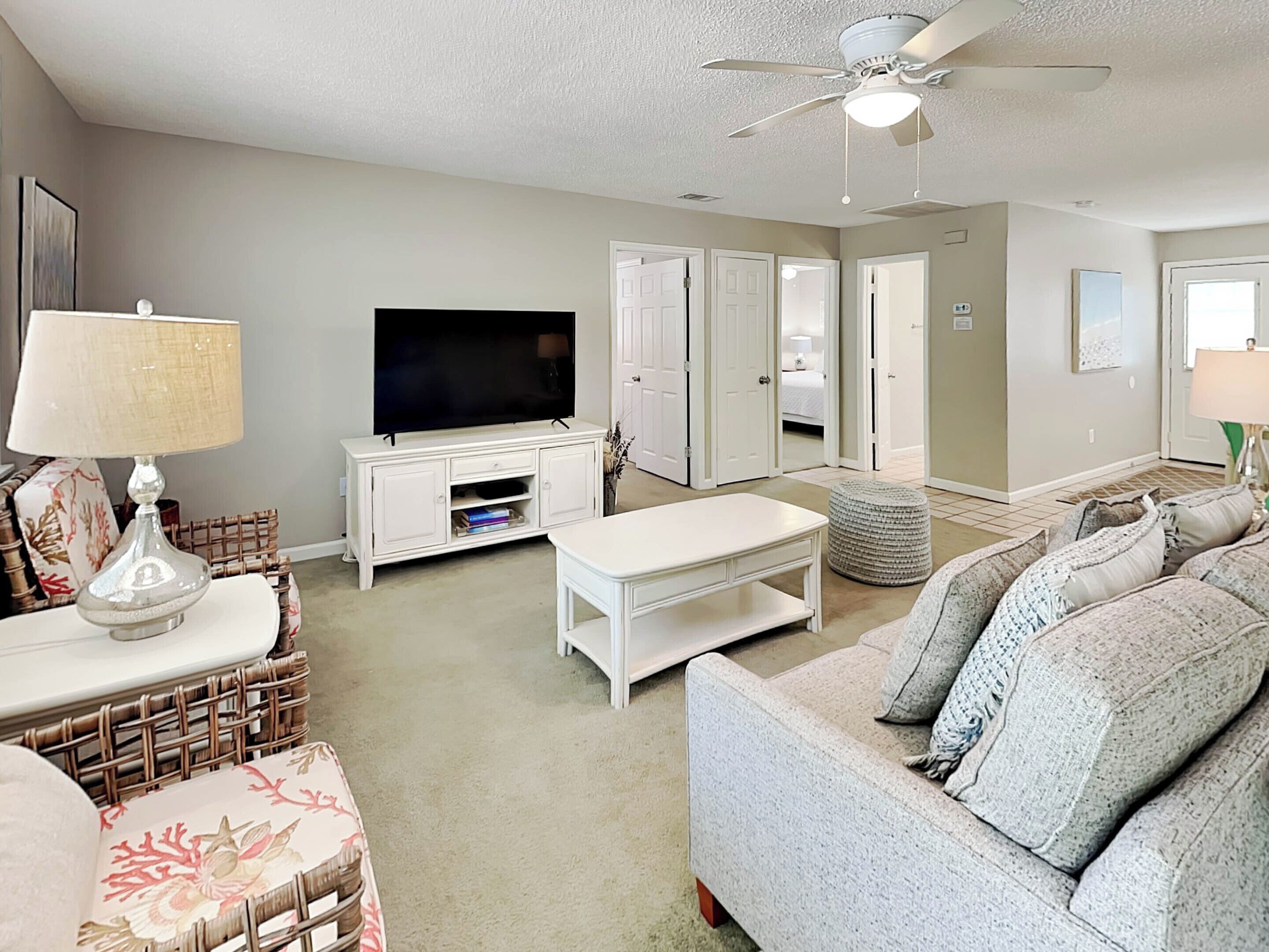 A living room with a flat-screen TV, white furniture, a ceiling fan, and a large window.