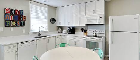 Una cocina compacta con armarios blancos, un refrigerador blanco, una estufa y un lavamanos. Hay una mesa redonda con cuatro sillas y un reloj en la pared.