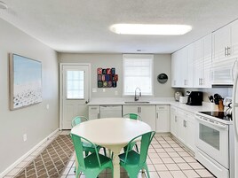 Una cocina con armarios blancos, una mesa blanca y sillas verdes. Se ve una ventana con persianas, un fregadero y una estufa.