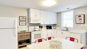 A compact kitchen with white cabinets, a stove, a microwave, and a sink. There is a dining table with red chairs and a decorative bowl on it. - Latitude Adjustment B Tybee Vacation Rentals (Tybee Island)