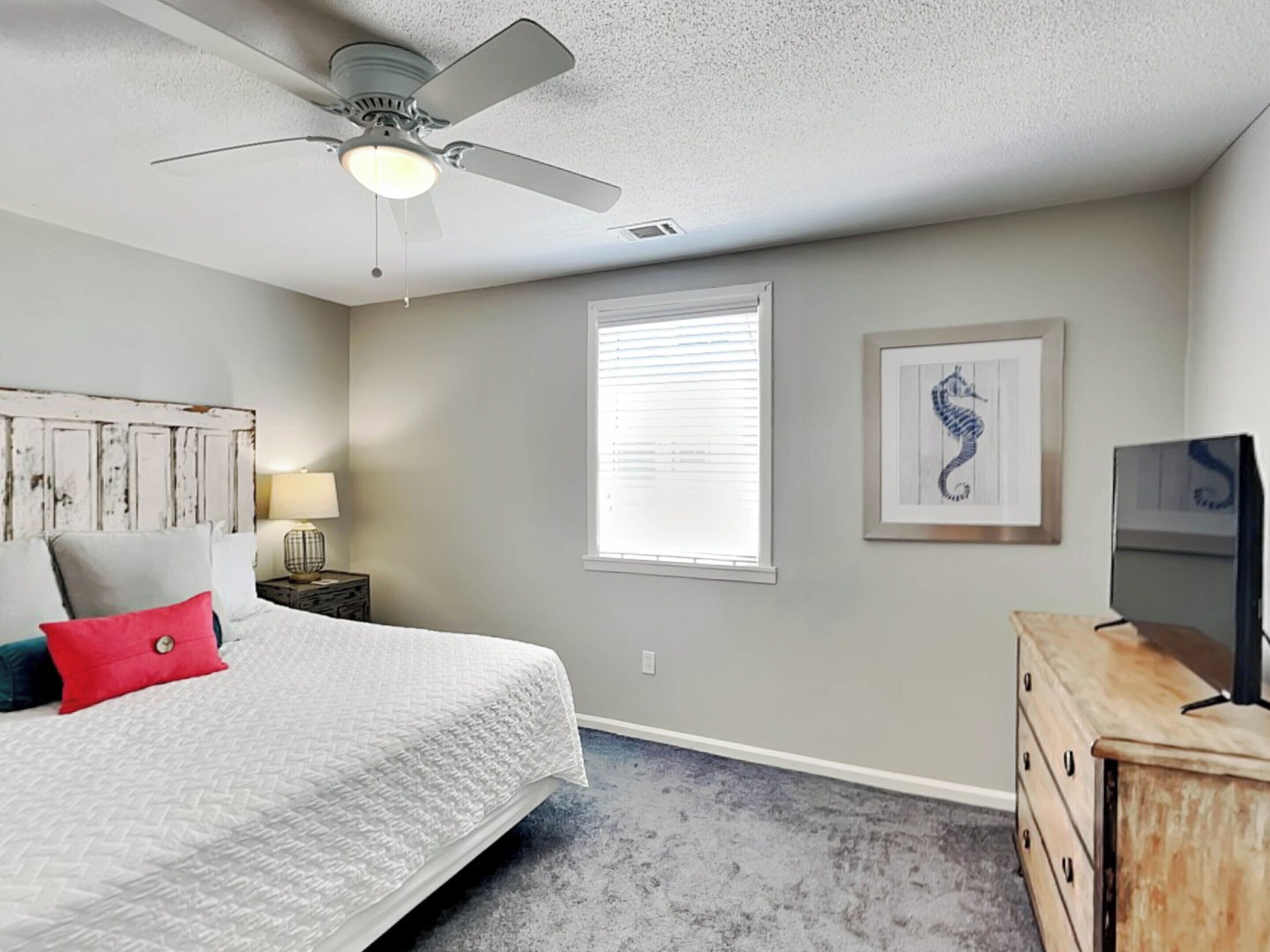 A bedroom with a bed, a ceiling fan, a window with blinds, a dresser, and a framed picture on the wall.