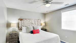 A bedroom with a large bed, two bedside tables with lamps, a ceiling fan, and a window with blinds. - Latitude Adjustment B Tybee Vacation Rentals (Tybee Island)