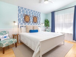 Un dormitorio con cama, una silla, una mesita de noche, una pared con diseño de hojas y una ventana con cortinas.
