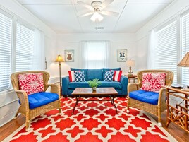 Uma sala de estar com um sofá azul, duas cadeiras de vime, um tapete com padrão vermelho e branco, e um ventilador de teto.