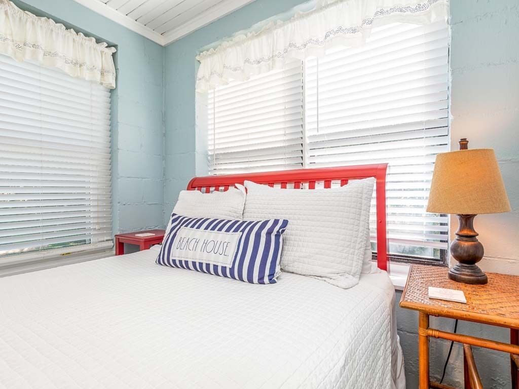 A bedroom with a bed, white bedding, a red headboard, a striped pillow, a lamp, and a small table.