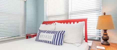 A bedroom with a bed, white bedding, a red headboard, a striped pillow, a lamp, and a small table.