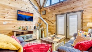 L’intérieur chaleureux d’un chalet, avec un revêtement mural en bois, un téléviseur fixé au mur, un canapé, une table basse et un meuble contenant des objets décoratifs.