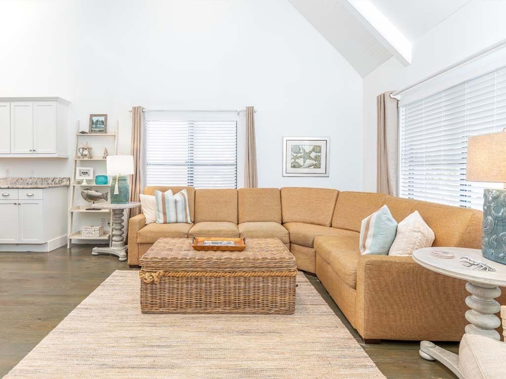 A living room with a sectional sofa, a wicker ottoman, a round coffee table, and a kitchen in the background.