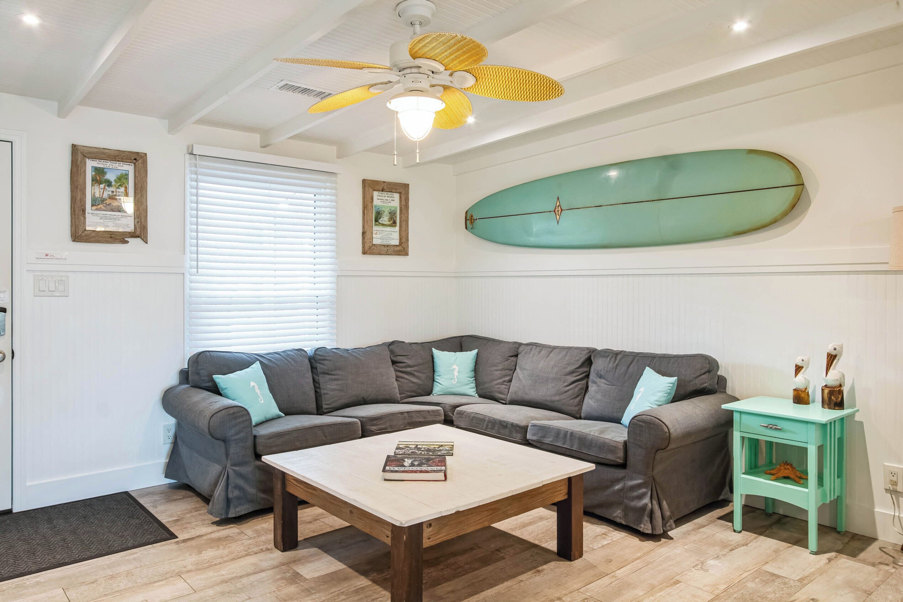 A living room with a gray sectional sofa, a wooden coffee table, a teal surfboard on the wall, and a small teal side table with decorative items.