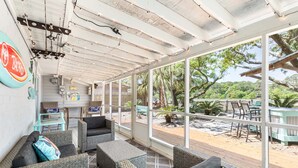 A covered outdoor seating area with wicker furniture and large windows overlooking a landscaped garden. - Lucky Charm by Tybee Vacation Rentals (Tybee Island)
