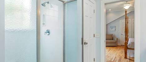 A modern bathroom with a glass shower enclosure, a white tiled floor, and a visible door leading to a living area with a wooden floor and a ceiling fan.