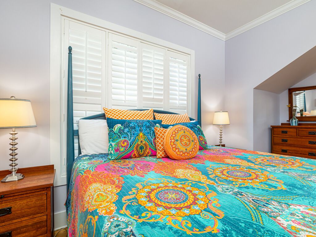 A bedroom with a bed covered in a colorful patterned bedspread, a window with white shutters, and a wooden dresser.