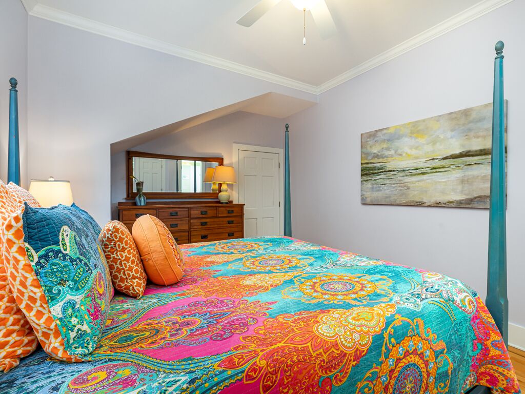 A bedroom with a colorful bedspread, a dresser, a mirror, and a painting on the wall.