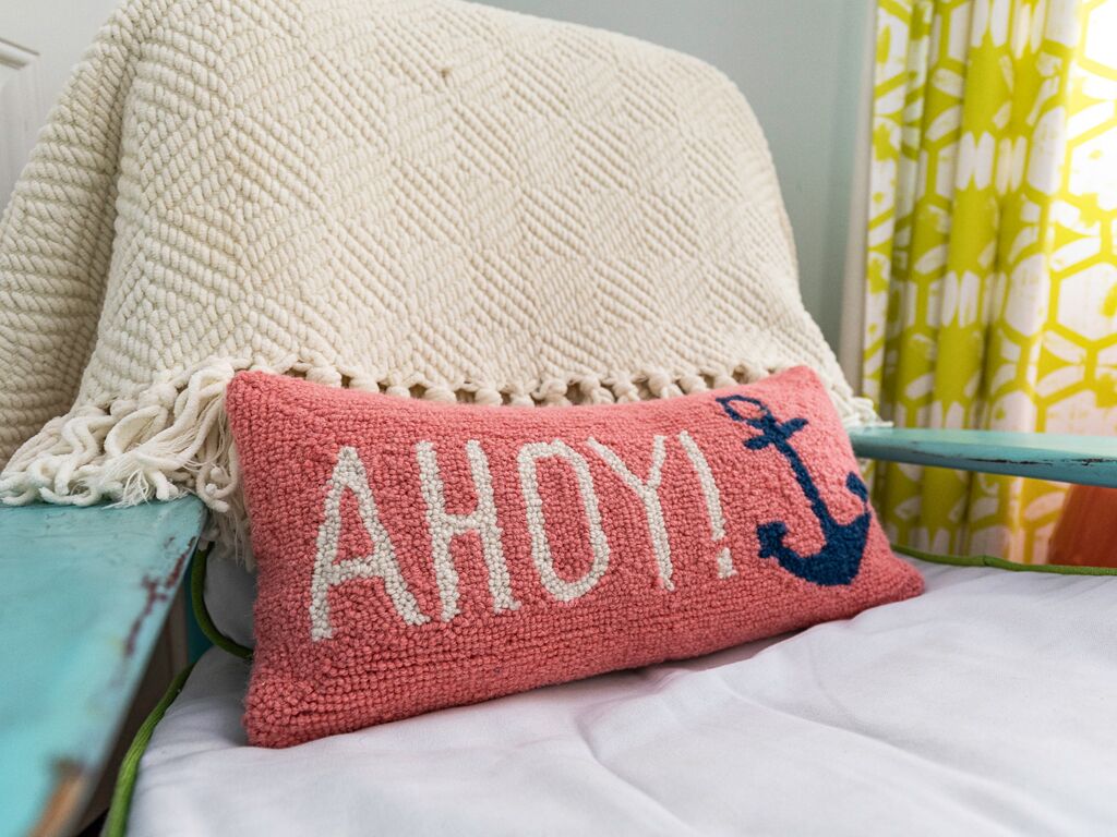 A cozy bed with a white knitted blanket and a pink pillow with embroidered text and a blue anchor.