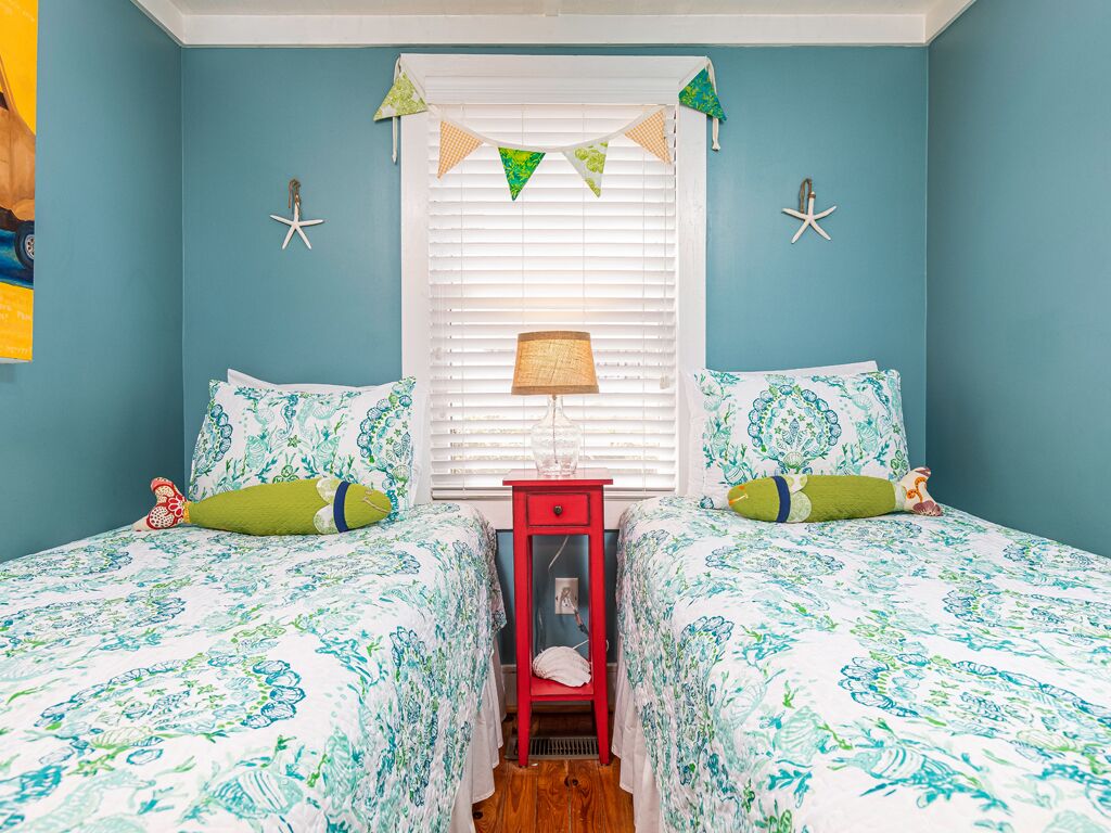 A room with teal walls, two beds with matching floral bedding, a red nightstand, a window with blinds, and starfish decorations.