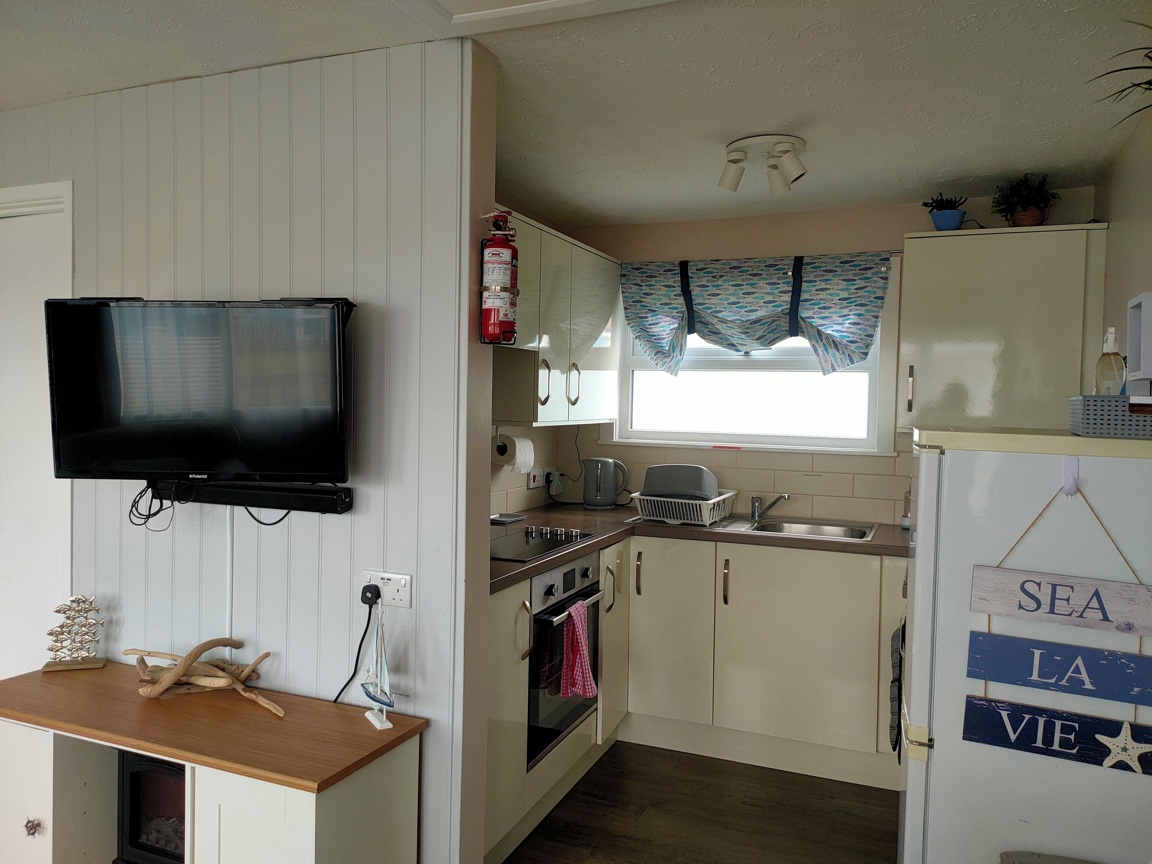 A compact kitchen with a flat-screen TV, a built-in oven, a sink, and a window with patterned curtains.