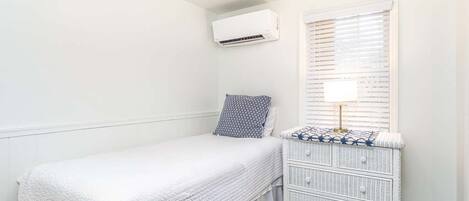 A small, neatly arranged bedroom with a single bed, a dresser, and an air conditioning unit on the wall.