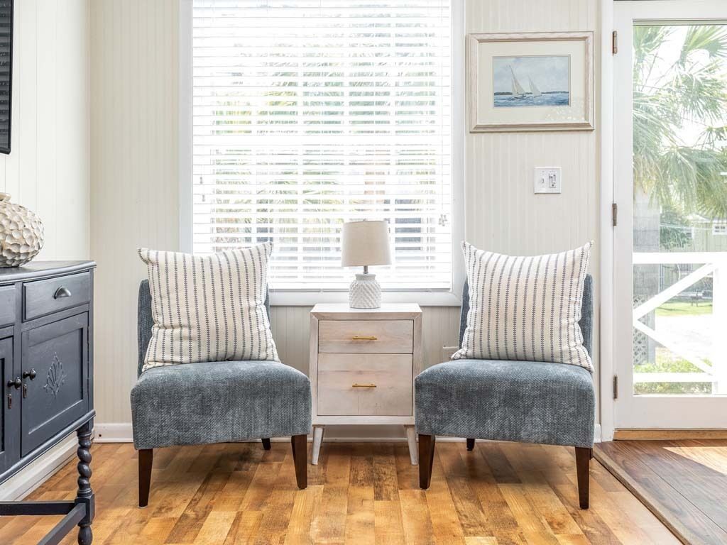 A room with two grey upholstered chairs with striped pillows, a white side table with a lamp, and a window with blinds.