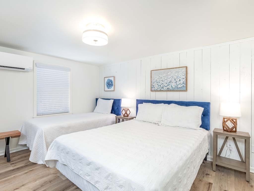 Une chambre avec deux lits, du linge de lit blanc, un plancher en bois, un luminaire au plafond et un climatiseur fixé au mur.