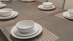 Une table à manger est dressée avec des bols et des assiettes blancs, disposés sur un plancher en bois texturé, avec un canapé aux tons neutres en arrière-plan.