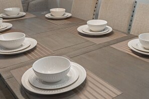 Une table à manger est dressée avec des bols et des assiettes blancs, disposés sur un plancher en bois texturé, avec un canapé aux tons neutres en arrière-plan.